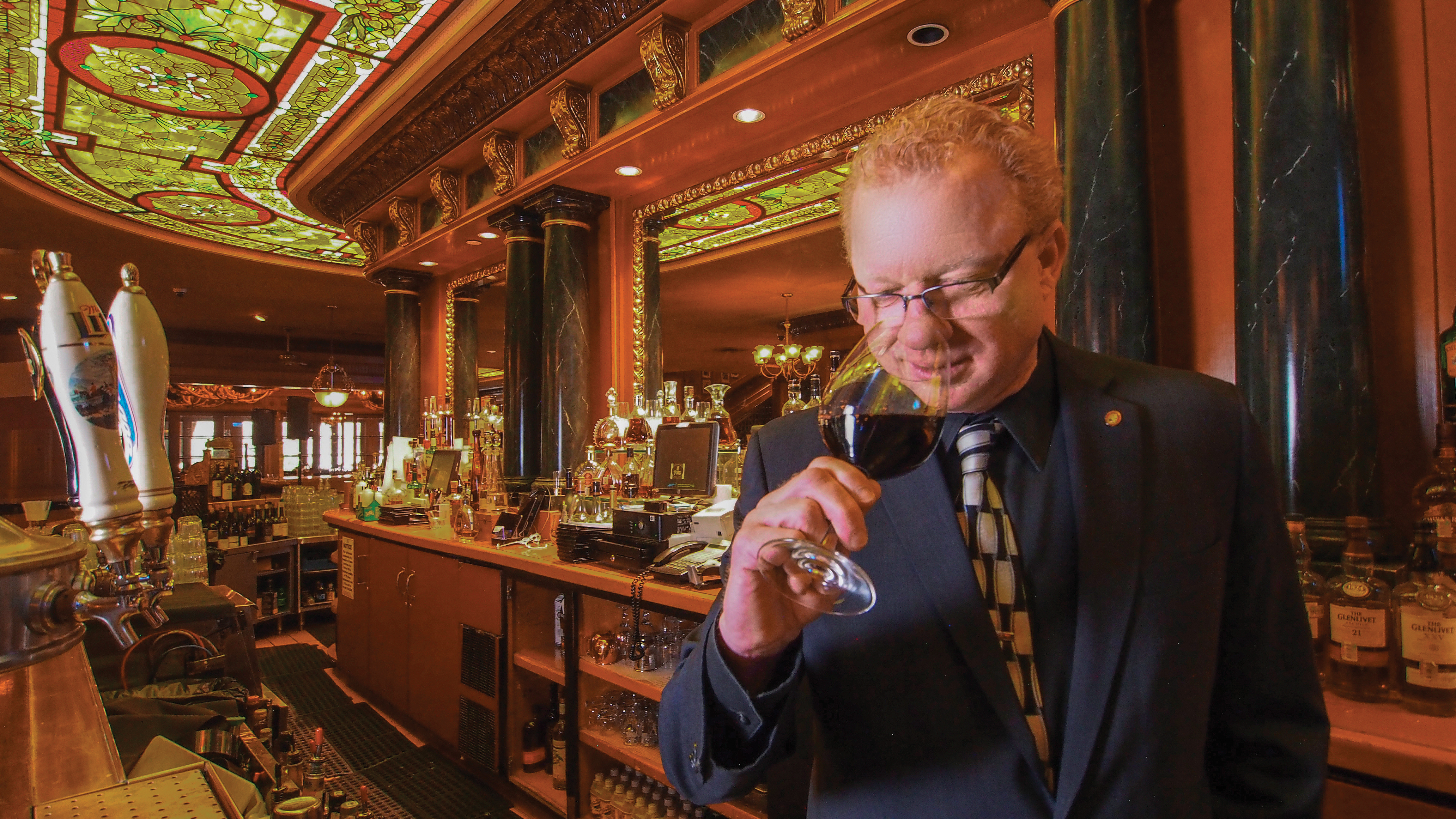 Dennis stands behind the bar and smells a glass of wine