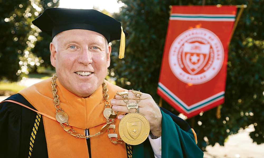 Benson, wearing regalia, holds his investiture medallion while standing in front of magnolia trees on the campus mall